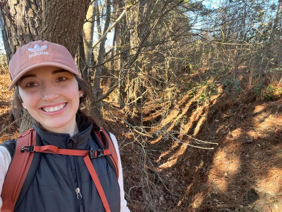 Mandi Taylor smiling beside a tree in bare winter woods, wearing a cap and a daypack, with pine needles covering the slope behind her.